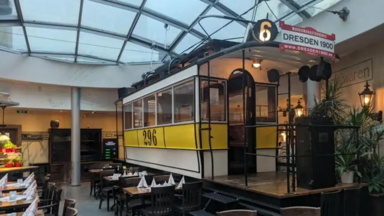 Eine historische Straßenbahn mit gelbem Streifen steht in einem Restaurant unter einem Glasdach, umgeben von gedeckten Tischen.