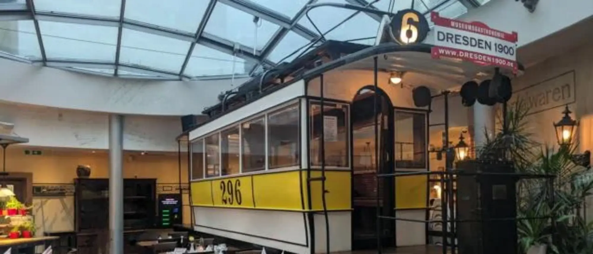 Innenaufnahme eines Restaurants mit einer historischen Straßenbahn als zentrales Dekorationselement. Die Straßenbahn ist gelb-weiß und trägt die Nummer 296. Darüber hängt ein Schild mit der Aufschrift "Dresden 1900". Um die Straßenbahn herum sind Tische und Stühle angeordnet, die für Gäste eingedeckt sind. Das Restaurant befindet sich unter einem Glasdach, das für viel natürliches Licht sorgt. Die Atmosphäre wirkt nostalgisch und gemütlich.