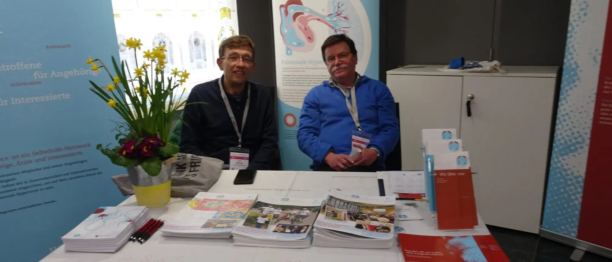 Das Bild zeigt Peter Bönning und Klaus Konz an einem gut gelegenen Infostand. Vor ihnen liegen verschiedene Informationsmaterialien, Broschüren und Flyer auf dem Tisch. 