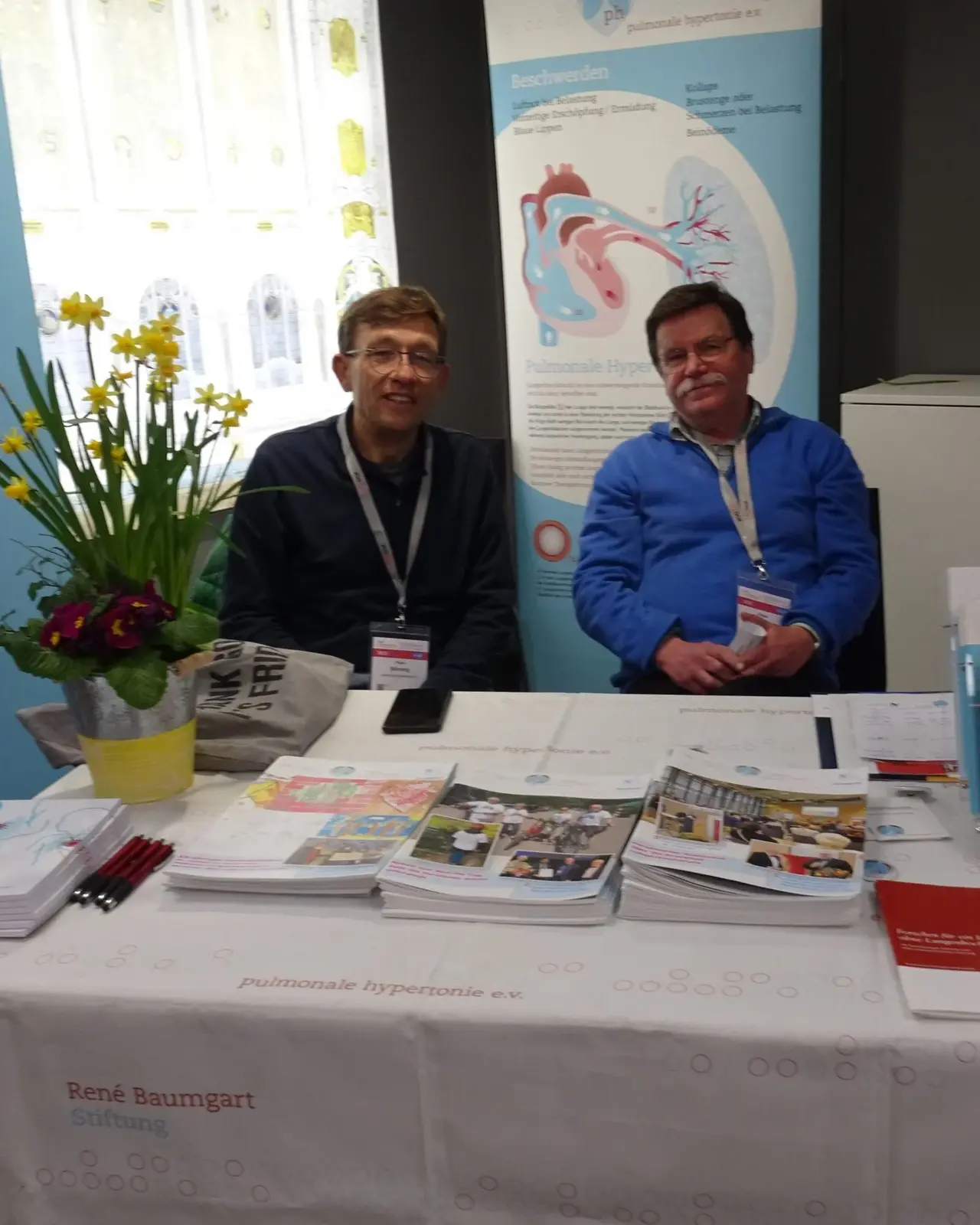 Das Bild zeigt Peter Bönning und Klaus Konz an einem gut gelegenen Infostand. Vor ihnen liegen verschiedene Informationsmaterialien, Broschüren und Flyer auf dem Tisch. 