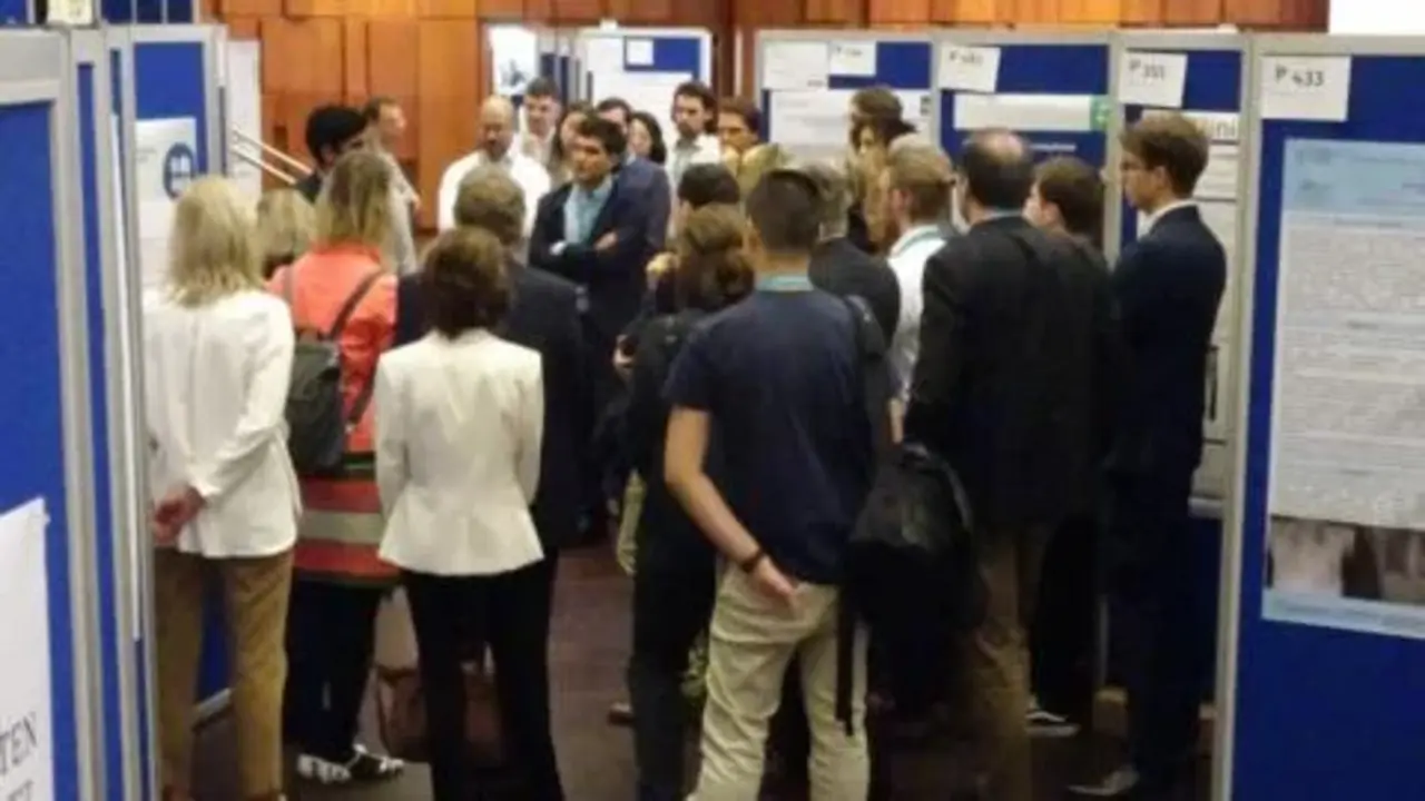 Eine Gruppe von Menschen versammelt sich in einer Ausstellungshalle vor mehreren blauen Posterwänden, auf denen wissenschaftliche Poster oder Präsentationen angebracht sind. Die Umgebung ist modern, mit Holzverkleidungen an den Wänden. Die Teilnehmer hören aufmerksam einer Präsentation zu.