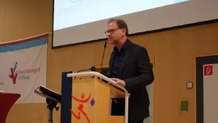 Dr. med. Matthias Held steht an einem Rednerpult und hält einen Vortrag. Im Hintergrund sind eine Leinwand mit einer Grafik und ein Banner der René Baumgart Stiftung zu sehen.