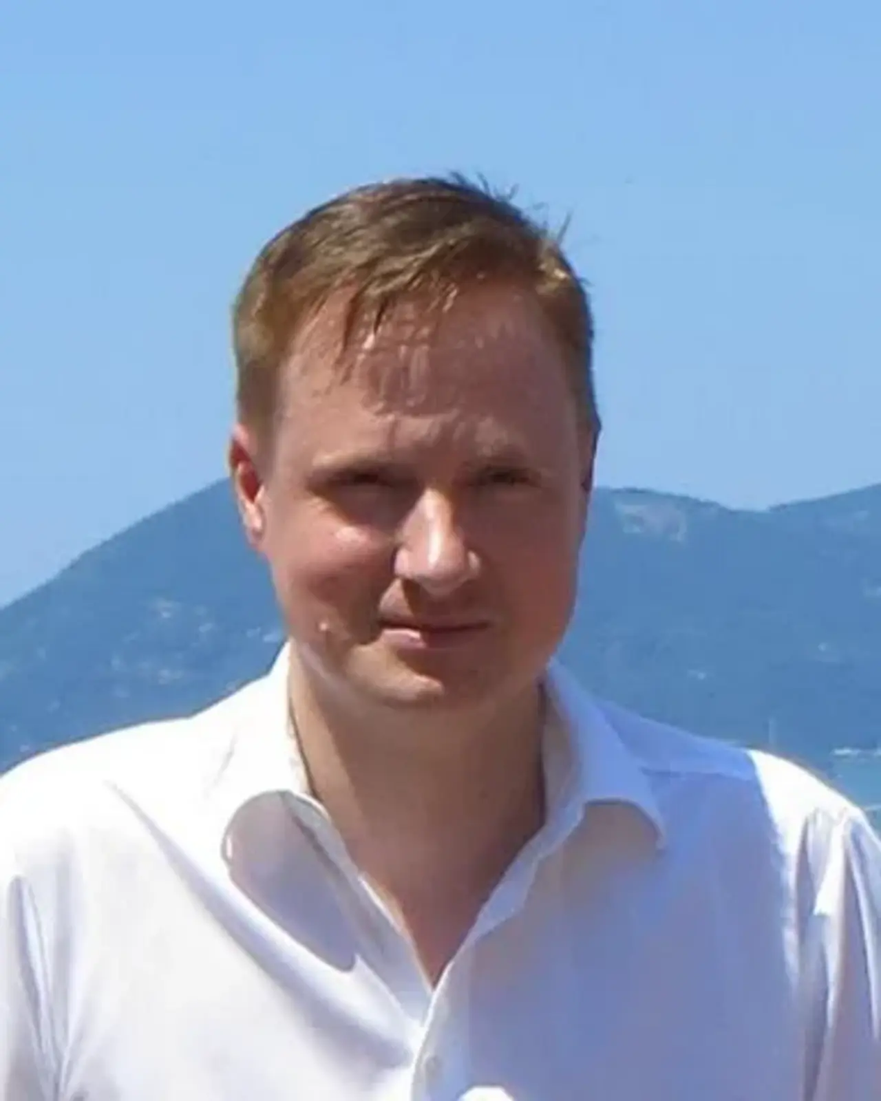 Roland Stenzel, ein Mann mit kurzem, blondem Haar, der ein weißes Hemd trägt. Er steht vor einer Kulisse mit Bergen und Meer im Hintergrund an einem sonnigen Tag.