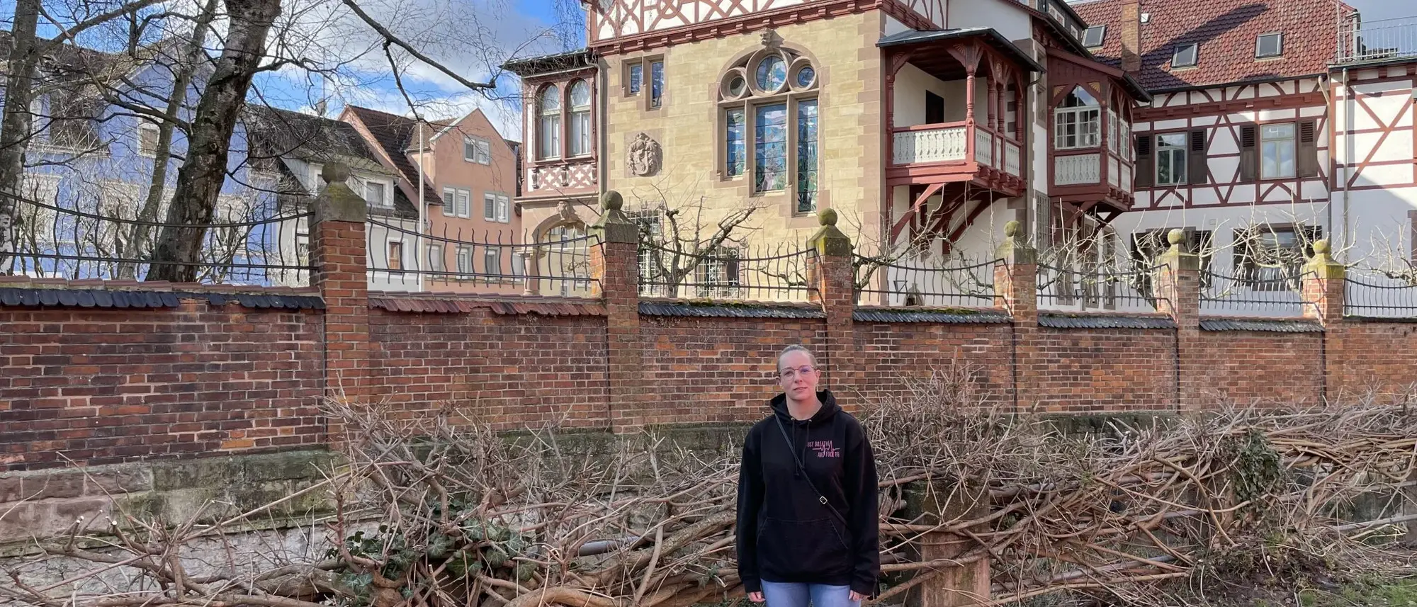 Das Bild zeigt Bianca bei einem Besuch in der Melanchthonstadt Bretten. Sie steht vor einer historischen Villa mit Fachwerkarchitektur, die hinter einer Backsteinmauer mit schmiedeeisernem Zaun liegt. Der klare Himmel und die kahlen Bäume lassen auf eine kühle Jahreszeit schließen. Bianca trägt eine schwarze Kapuzenjacke und lächelt in die Kamera.