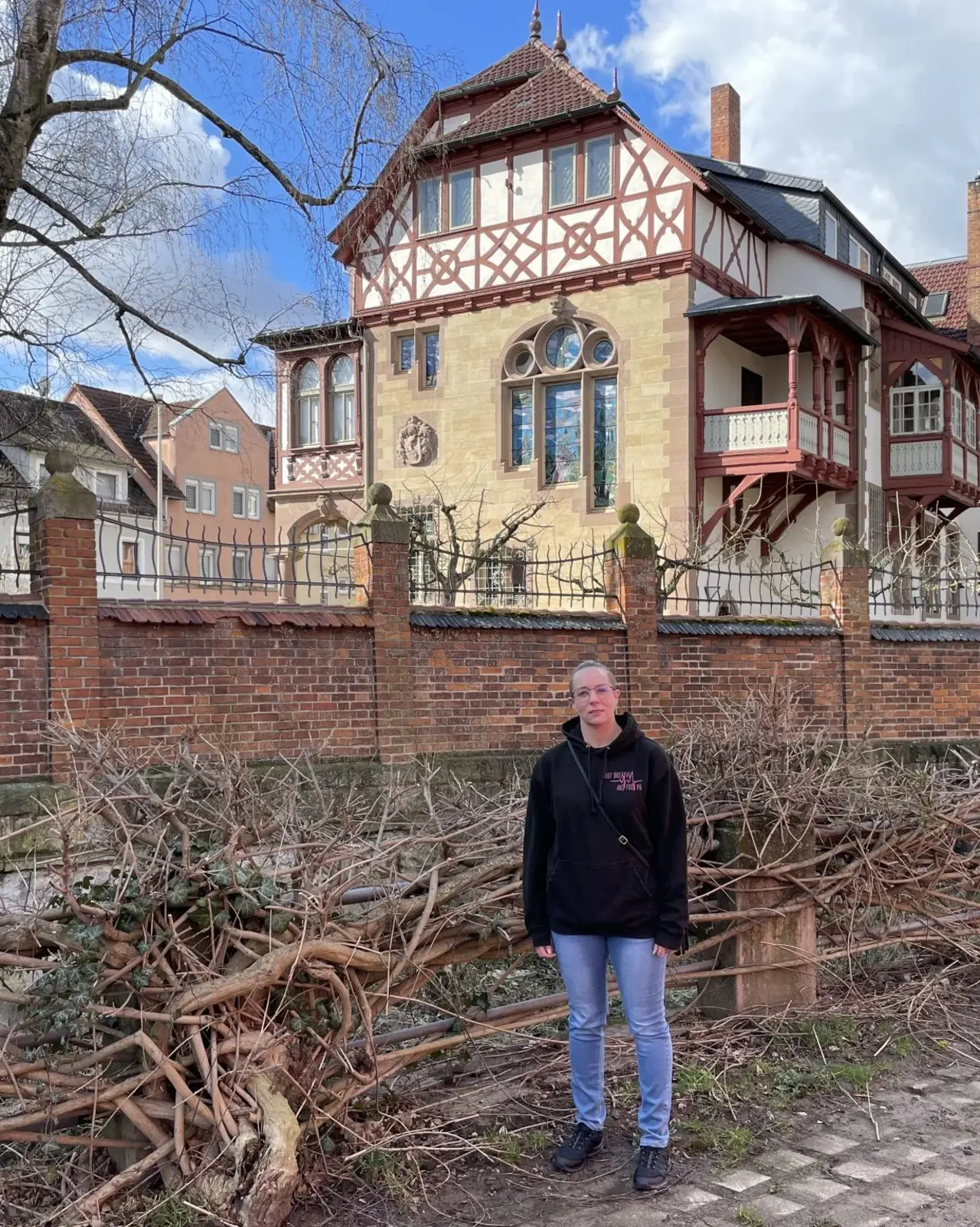 Das Bild zeigt Bianca bei einem Besuch in der Melanchthonstadt Bretten. Sie steht vor einer historischen Villa mit Fachwerkarchitektur, die hinter einer Backsteinmauer mit schmiedeeisernem Zaun liegt. Der klare Himmel und die kahlen Bäume lassen auf eine kühle Jahreszeit schließen. Bianca trägt eine schwarze Kapuzenjacke und lächelt in die Kamera.