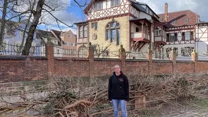 Das Bild zeigt Bianca bei einem Besuch in der Melanchthonstadt Bretten. Sie steht vor einer historischen Villa mit Fachwerkarchitektur, die hinter einer Backsteinmauer mit schmiedeeisernem Zaun liegt. Der klare Himmel und die kahlen Bäume lassen auf eine kühle Jahreszeit schließen. Bianca trägt eine schwarze Kapuzenjacke und lächelt in die Kamera.